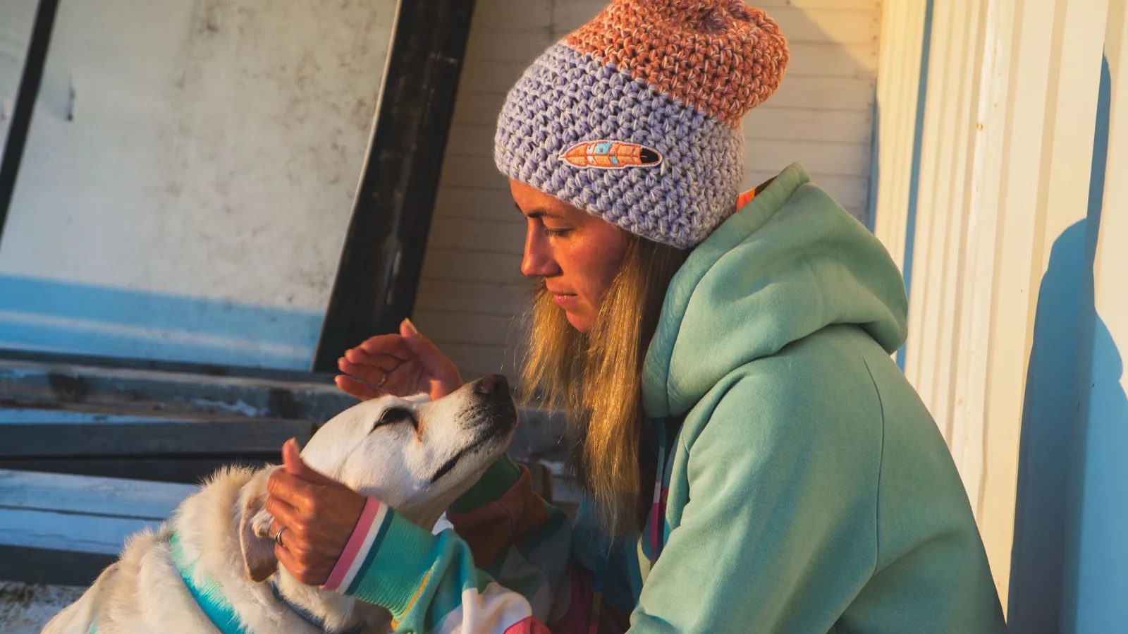 A woman in a hand-crocheted Polisze beanie crouches in golden evening light, holding a small dog dressed in a printed Polisze sleeve.