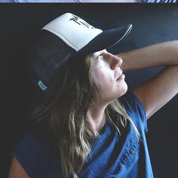 Woman in a white Polisze trucker cap and printed tee, lit against a dark background.